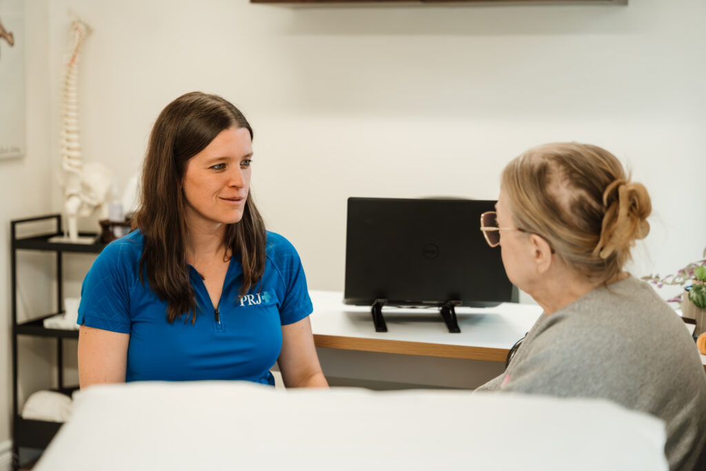 Deux femmes discutent assises dans un bureau de la clinique Physio PRJ de Repentigny, une avec un chandail bleu portant le logo de la clinique face à l'appareil, une plus âgée de dos, portant un chandail gris. 