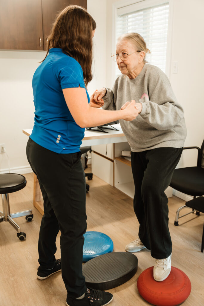 Une femme debout dans un chandail bleu et un pantalon noir, de dos, tient les mains d'une personne âgée debout devant elle et dont le pied gauche est déposé sur un coussin rond orange au sol. 